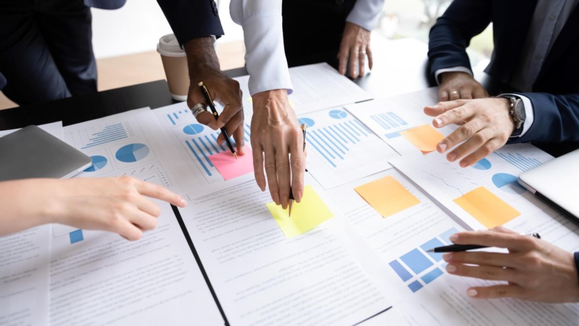 Close-up shot of a large table with documents spread out on it, with people pointing to several areas, collaborating on a report