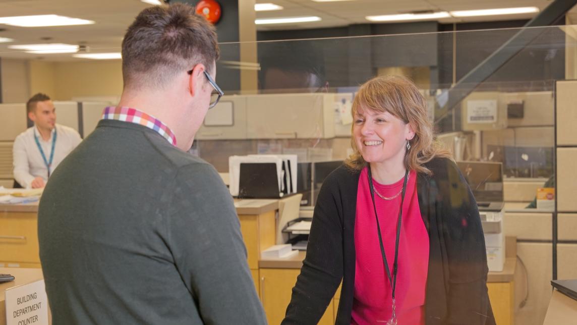 A woman smiles through a clear plastic barrier as she assists a man at the building department counter in City Hall.