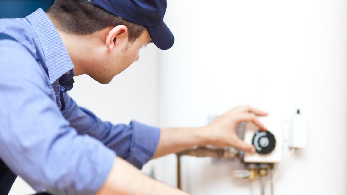 A man in a blue shirt and ballcap adjusts a dial on the side of a water heater.