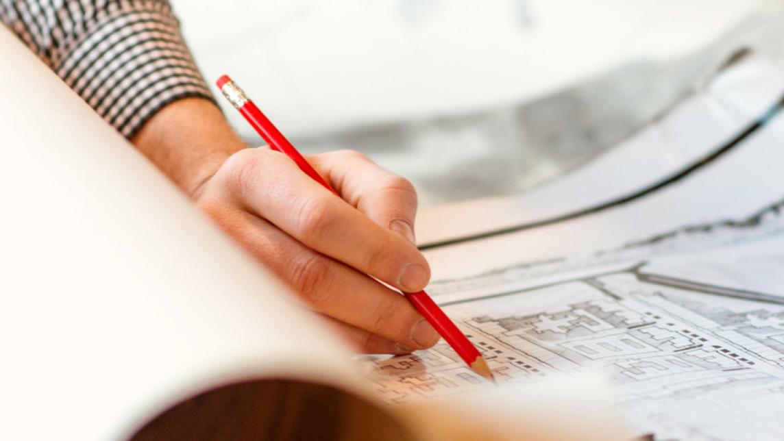 A person with a red pen spreads and marks now a note on a large paper plan.