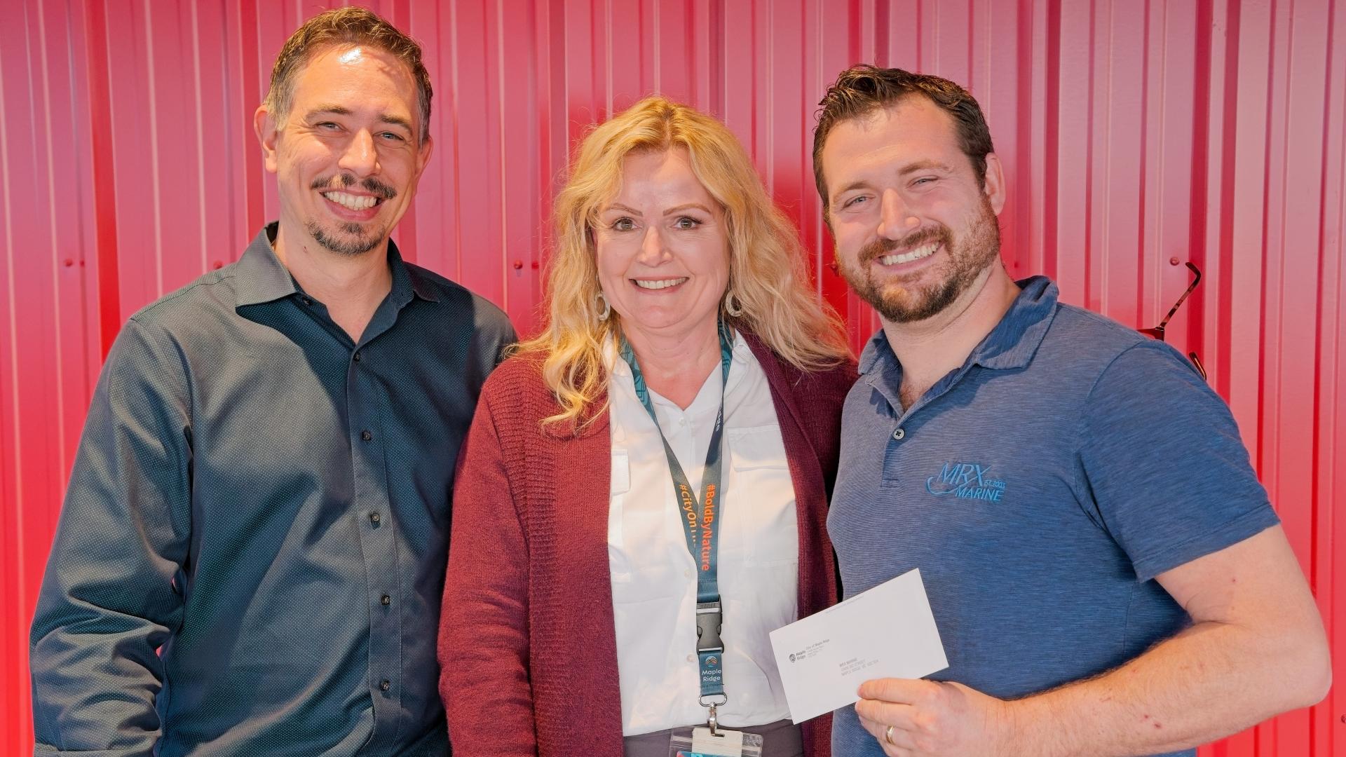 A man holds up a cheque and smiles next to the Masters of Ceremonies at the 2024 Innovation Awards Night.