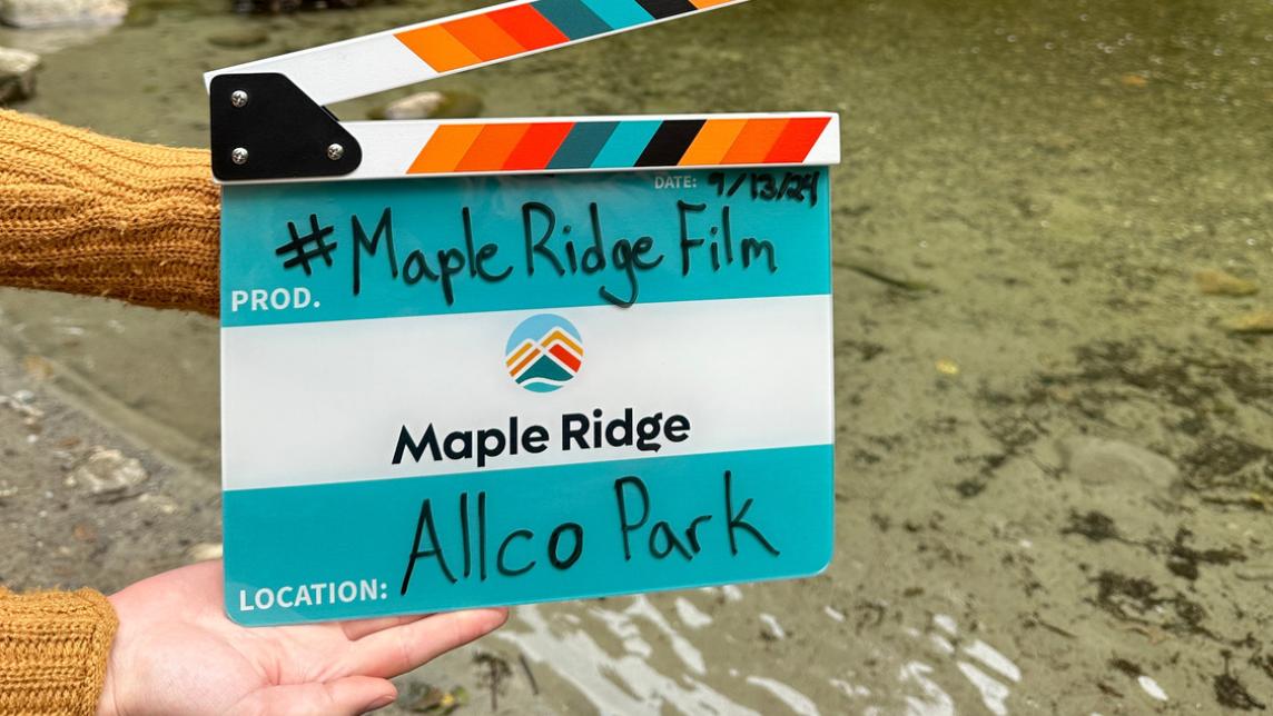 An offscreen woman holds out a City-branded film clapper with the words, #MapleRidgeFilm and Allco Park on it. The Maple Ridge logo sits in the middle.