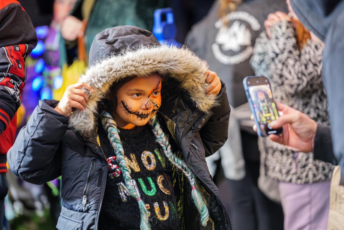 A child smiles and lifts the fur-lined hood of their jacket as their photo is taken with a cell phone.
