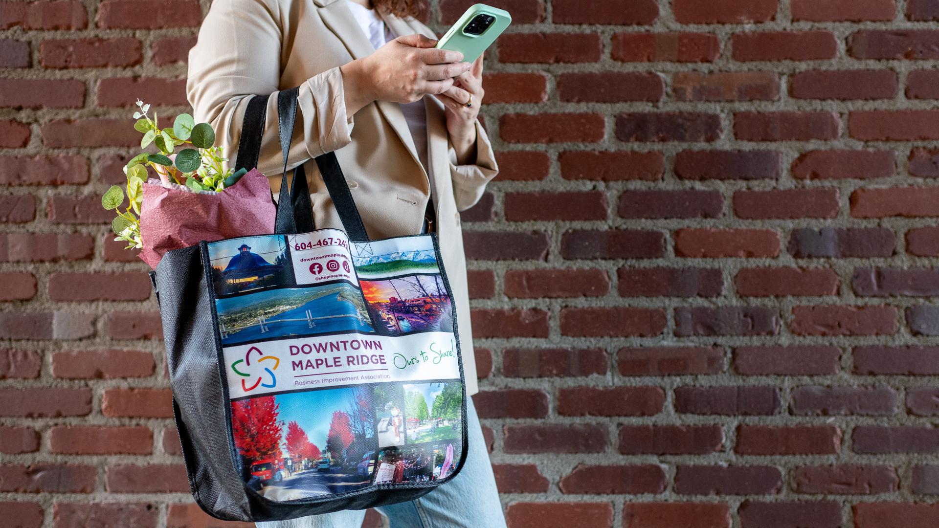 A woman wearing a beige jacket checks her phone while holding a shopping bag with groceries spilling out of the top.