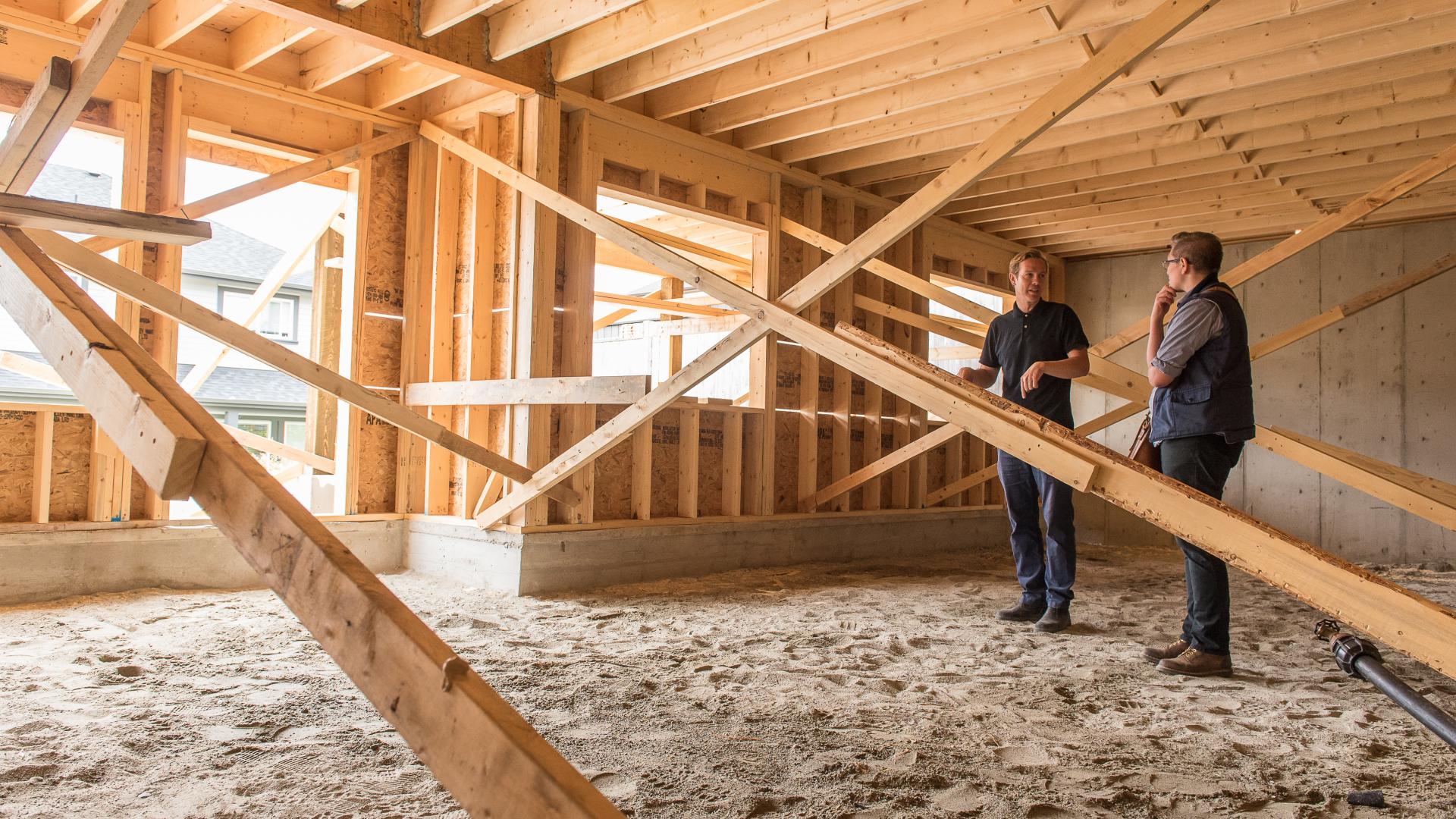 Two men in dark clothes stand inside a house under construction, having a discussion.