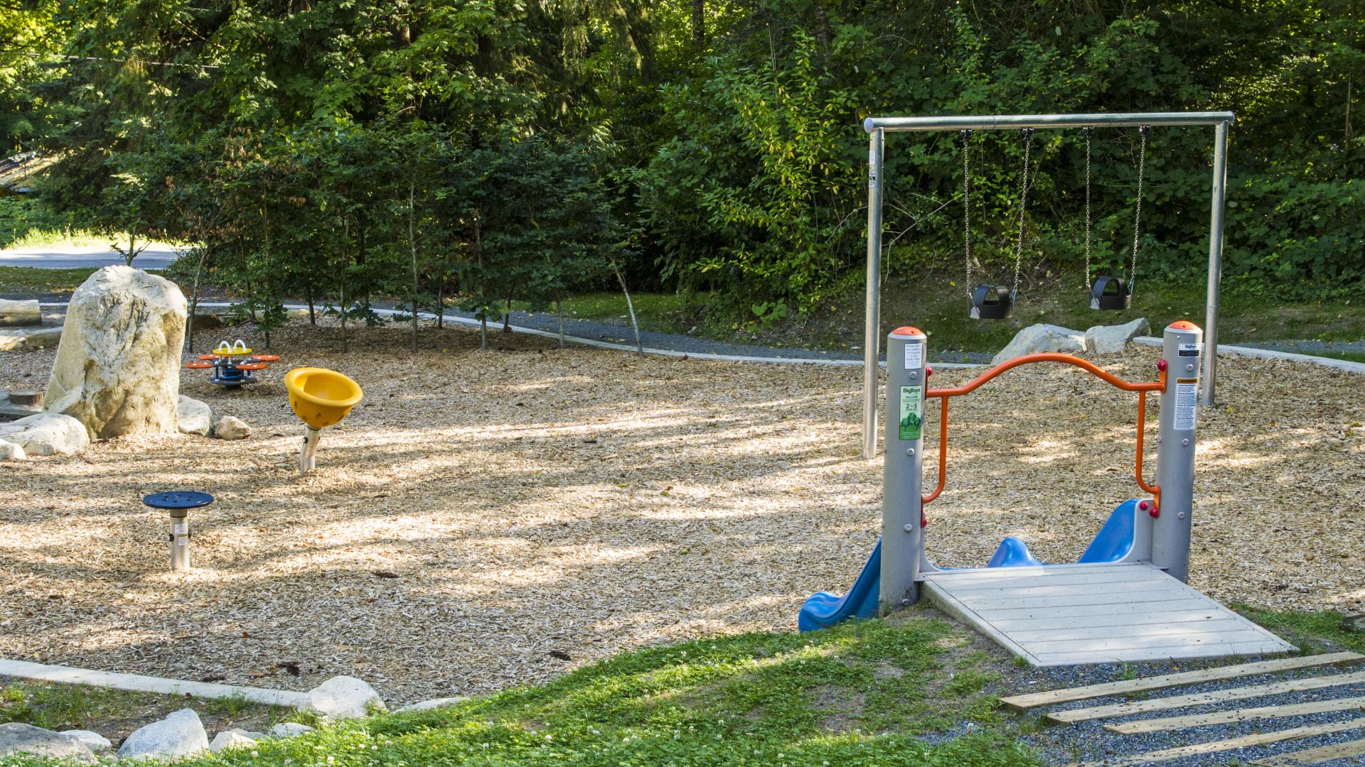 The variety of playground equipment is on a bed of mulch. There are swings and spinning structures, along with other equipment.