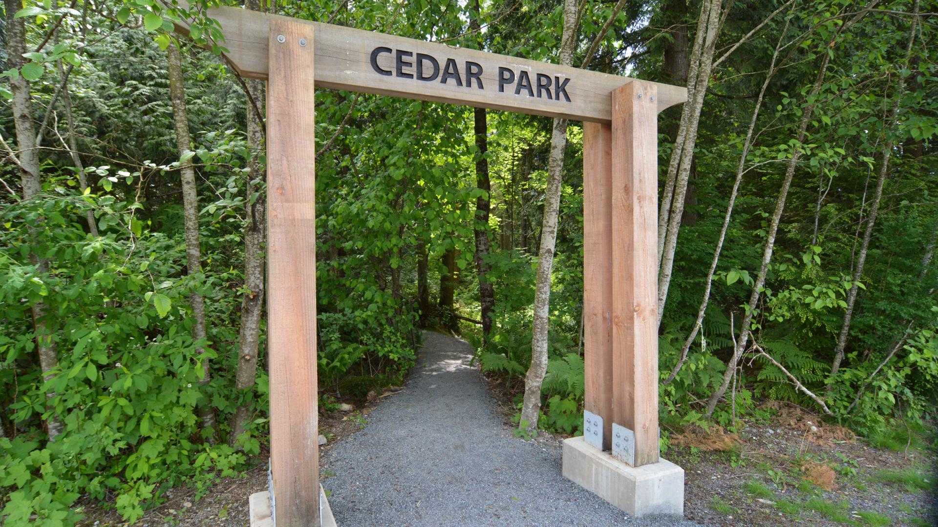 A wooden arch with Cedar Park on it over a paved pathway leading into a forest.