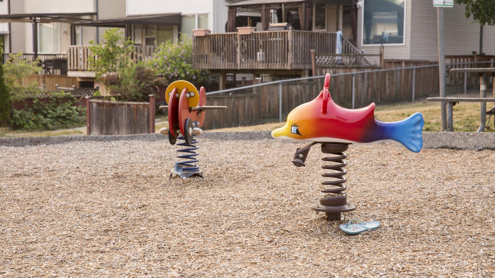 Two colourful metal spring rockers, one styled as a rainbow-coloured fish and the other as a red airplane sit in a bark mulch play area.