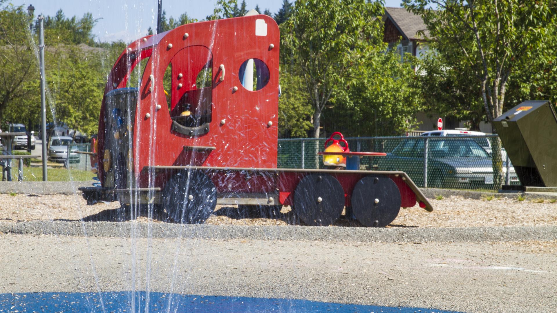 A fountain from the spray park splashes up water. Behind, there is a red, vehicle-like playground.