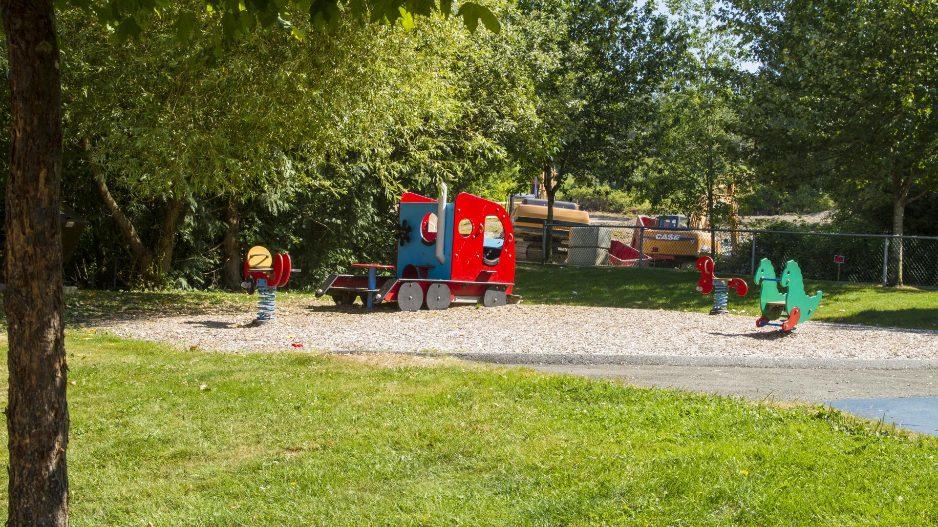 A grassy area surrounded by trees. At the centre, a sandy patch and a playground.