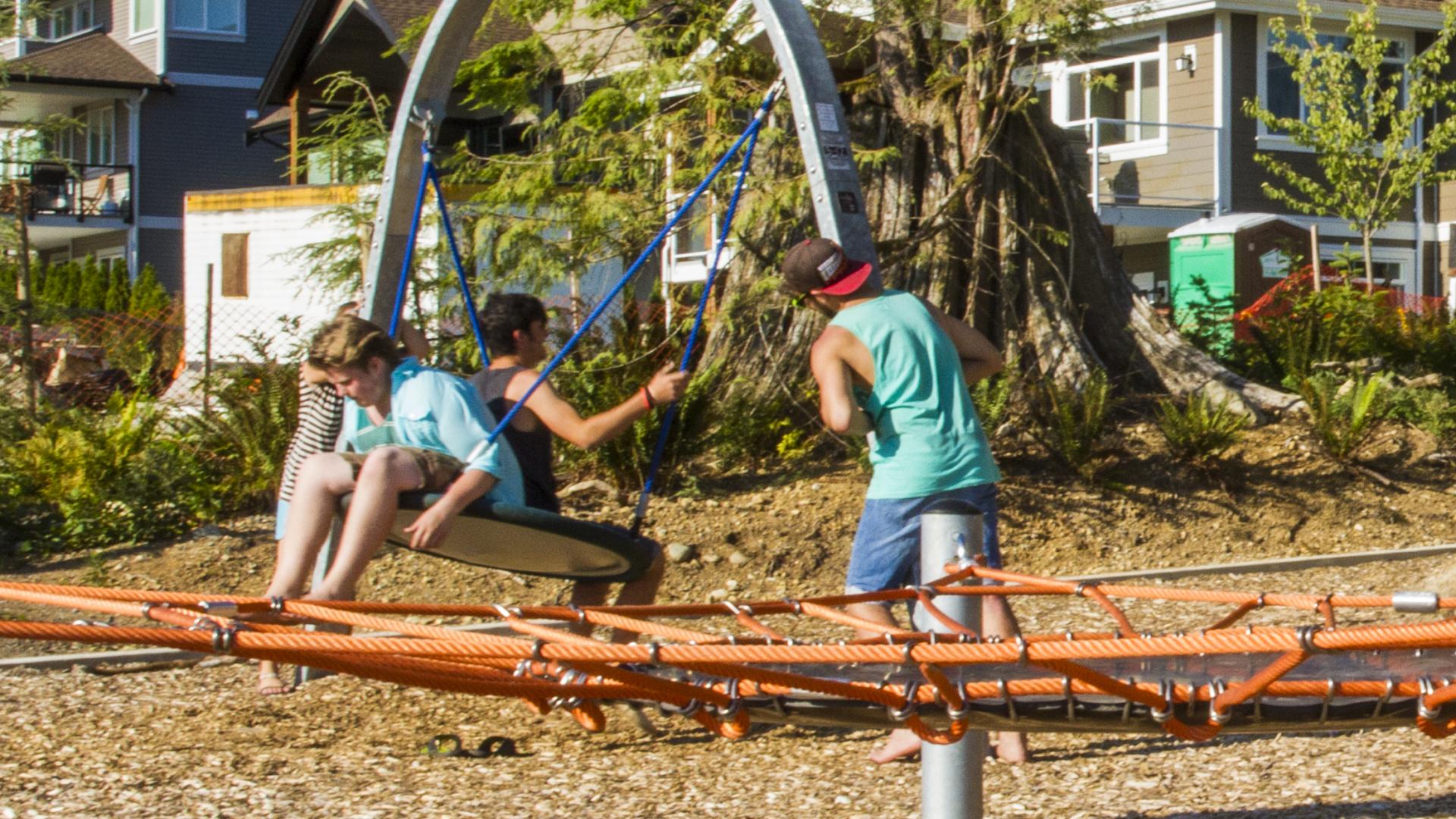 Two kids share a large circular swing, facing back to back. Another child watches. In the foreground, ropes from a rope climb are visible.