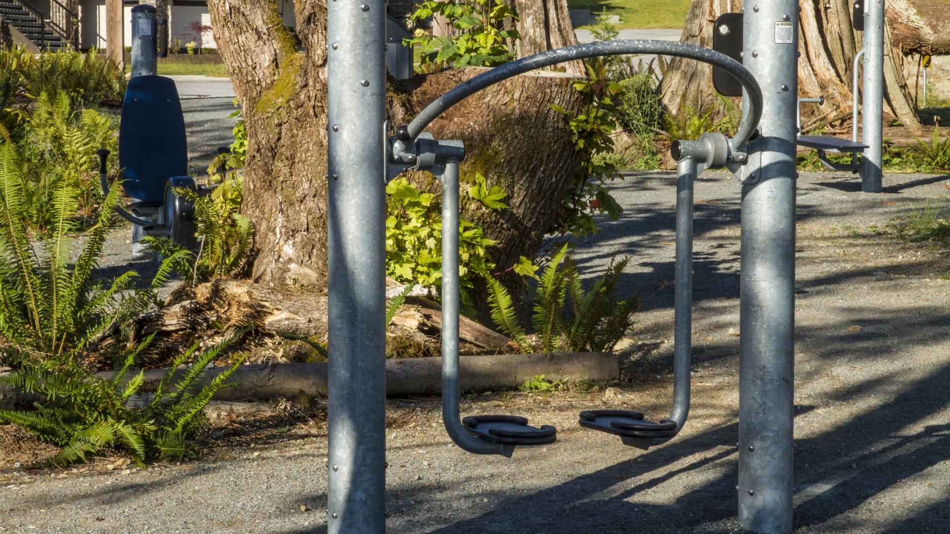 An manual elliptical machine. There is a paved area with a small garden in the middle and other manual work out equipment in the background.