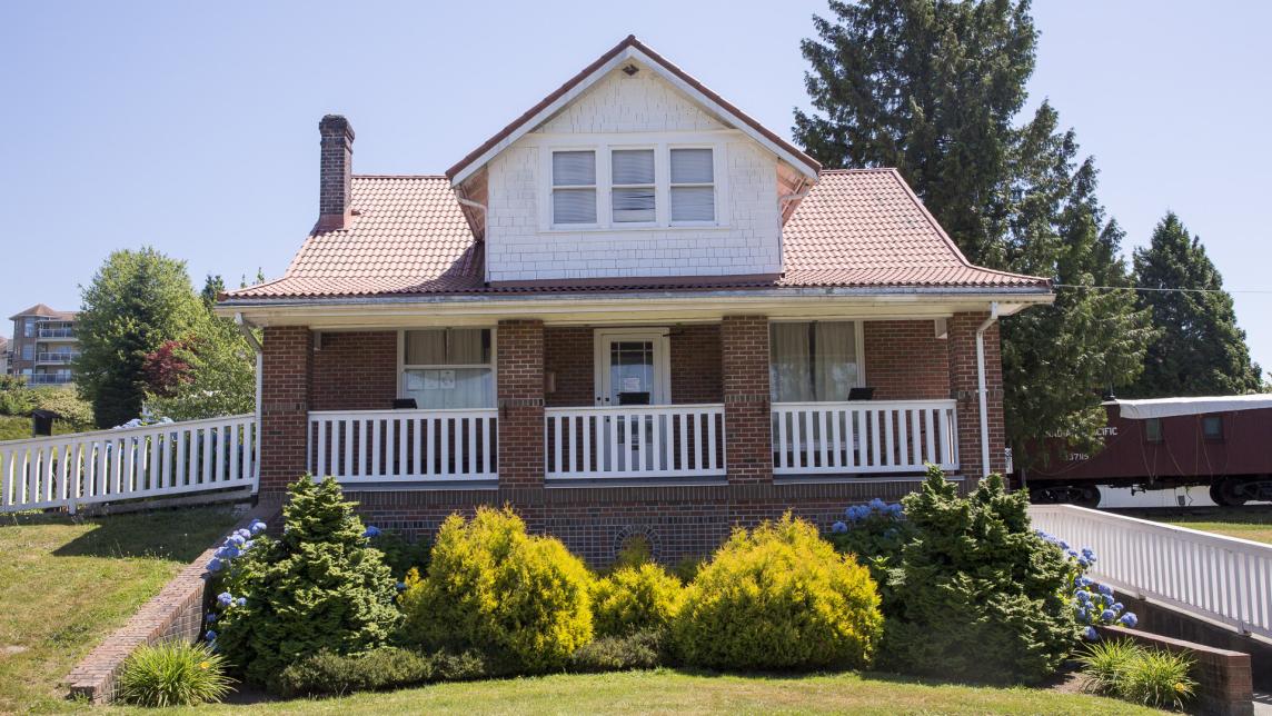 A large brick museum sits above a paved walkway lined with bushes next to a traincar.