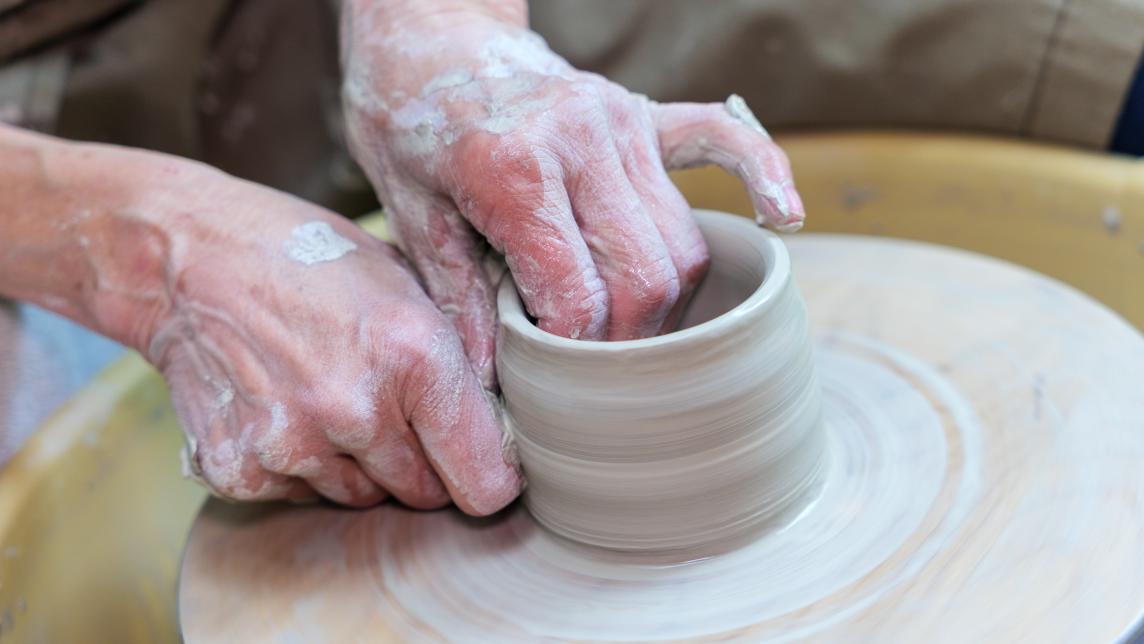 An artisan shapes clay into a cup-like vessel on a pottery wheel.