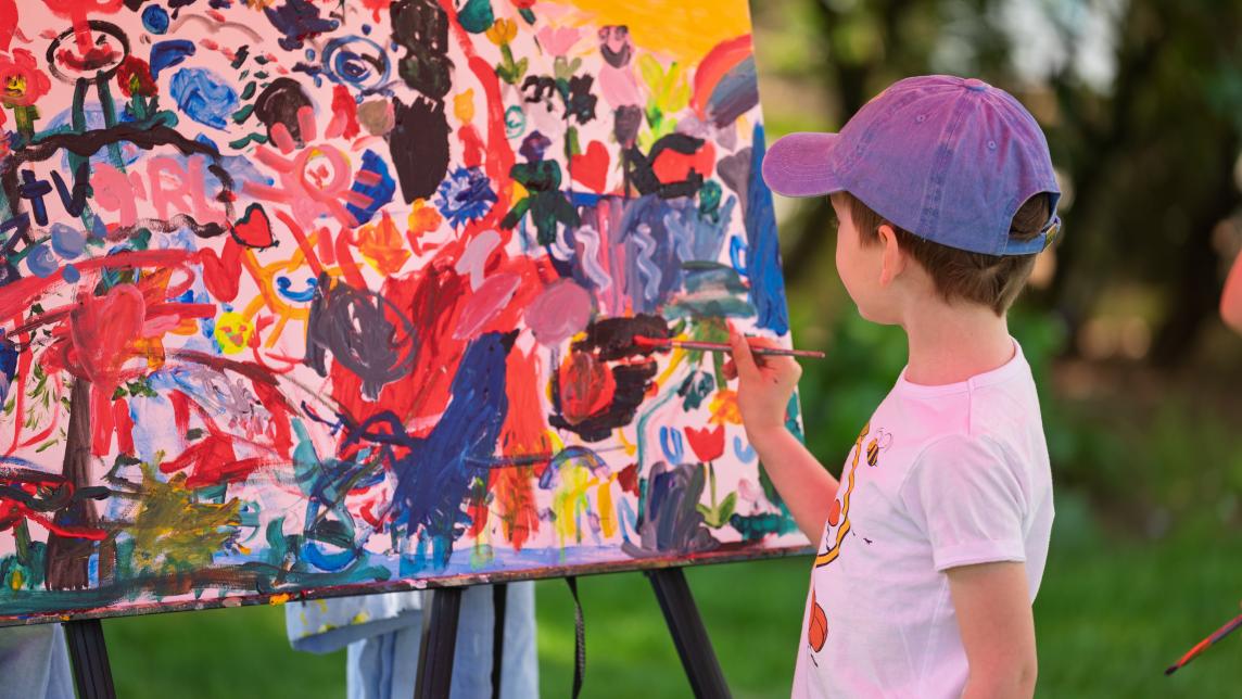 A young boy adds paint to a canvas outdoors.
