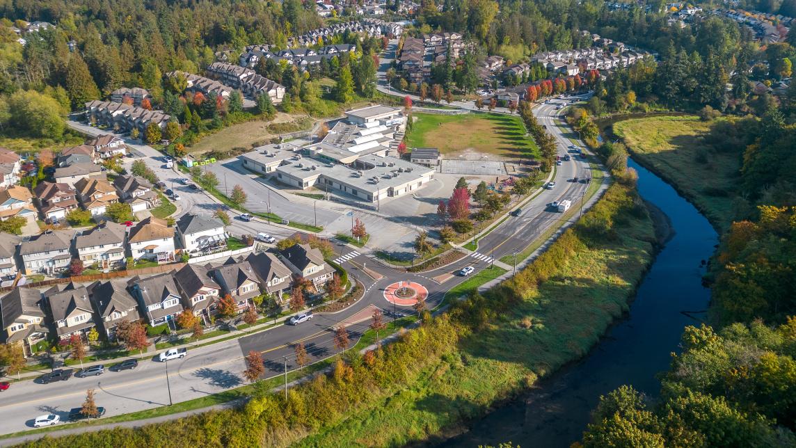 An aerial view of a road with a roundabout that hugs a river on one side, and homes on the other.