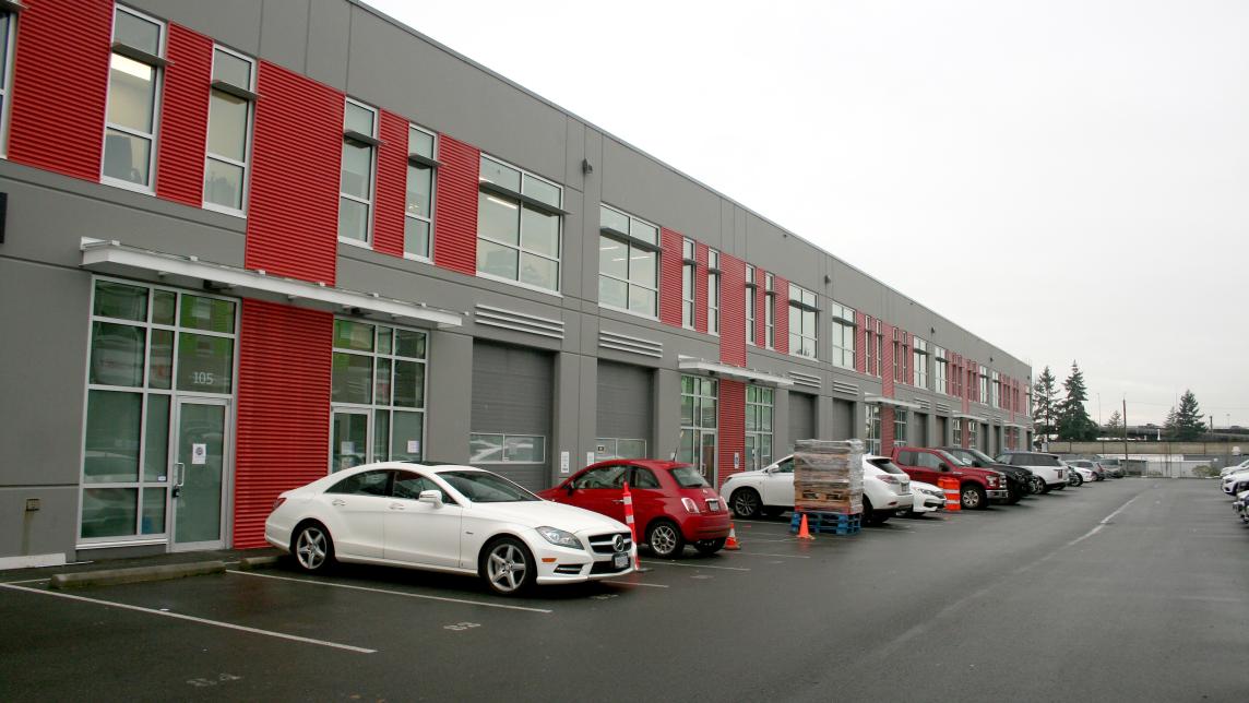 A line of cars parks right next to a long grey-and-red industrial building.