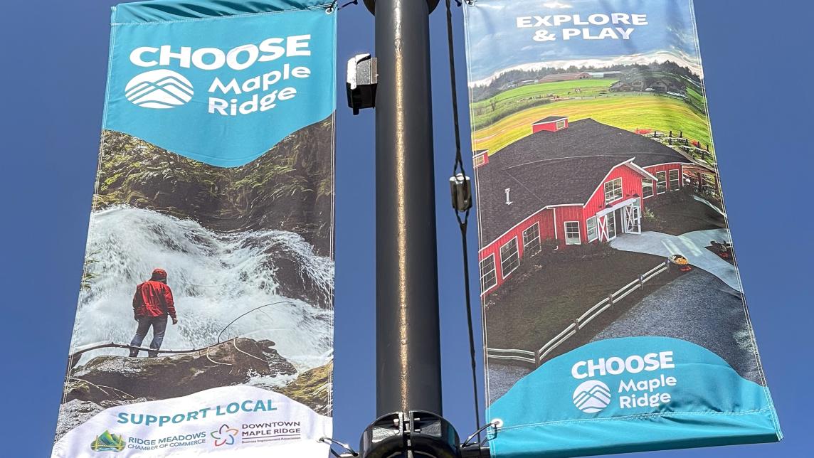 Two of the 2025 Choose Maple Ridge Street Banners, one showing a red farmhouse and the other a person in front of a waterfall.