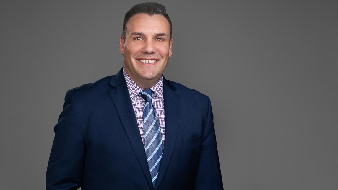 A man wearing a blue-toned suit with a striped tie smiles jovially.