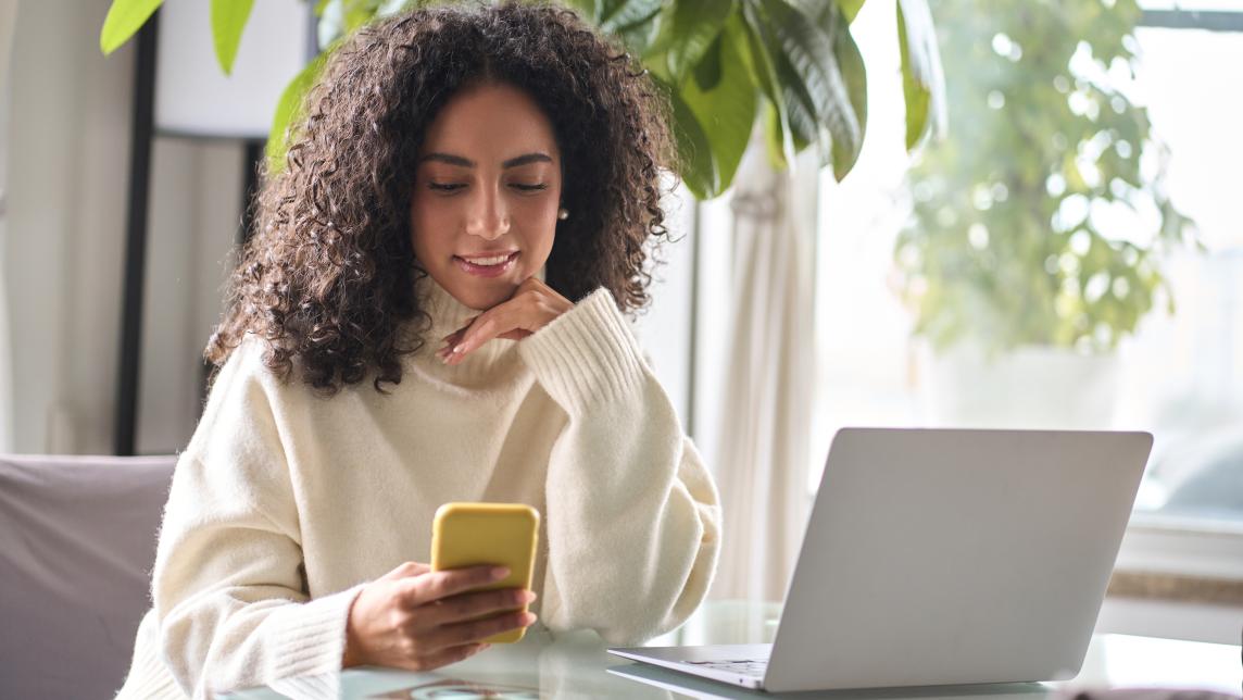 woman looking at her mobile phone sitting at a desk with a laptop on it