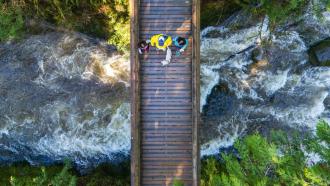 An aerial view of three people walking their dog across a wooden bridge overtop a raging river.