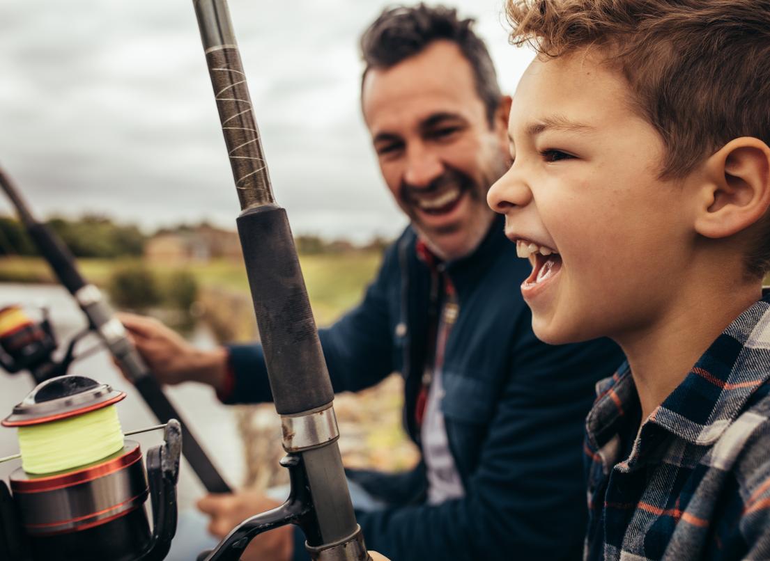 A young boy and his father fish together at the edge of a lake.