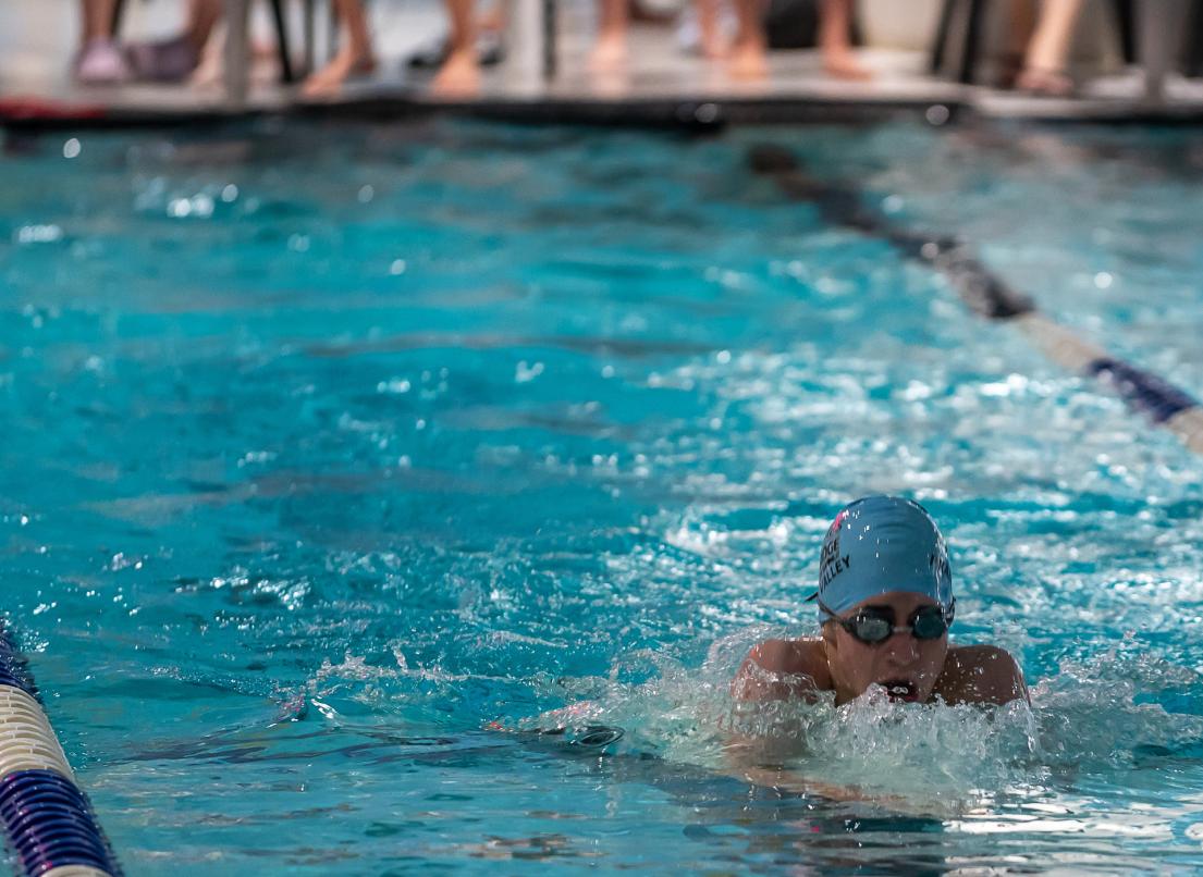 A racer surges through the water with onlookers crowded at the edge of the indoor pool.