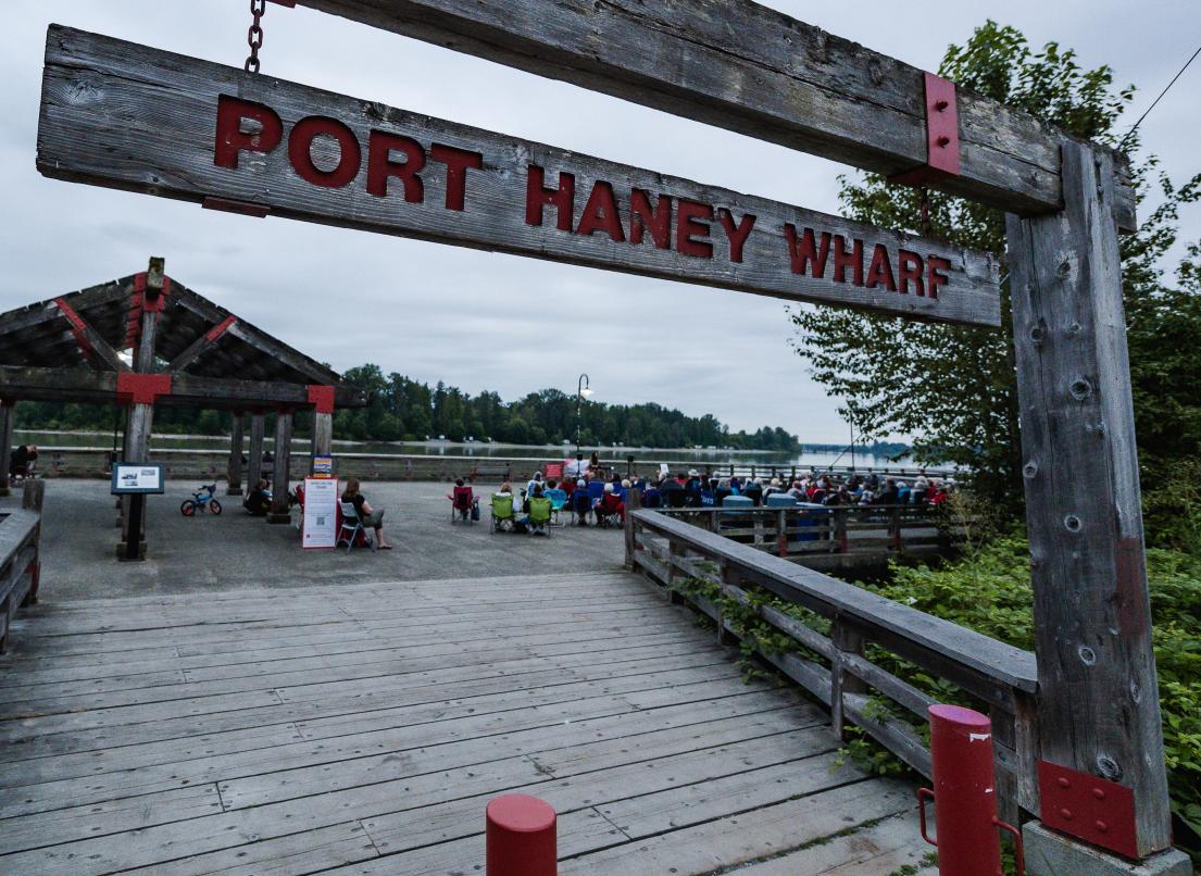 A wooden entranceway stands above the planks of a wharf where a live performance it being held. It proclaims "Port Haney Wharf" in red letters.