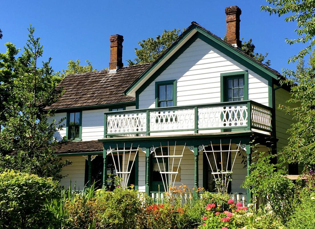 A white house with green trim is surrounded by green bushes, flowers and meticulously landscaped trees.