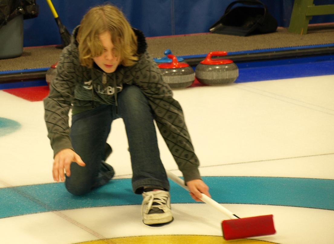 A young boy kneels dramatically on the ice as he lines up his next curling shot.
