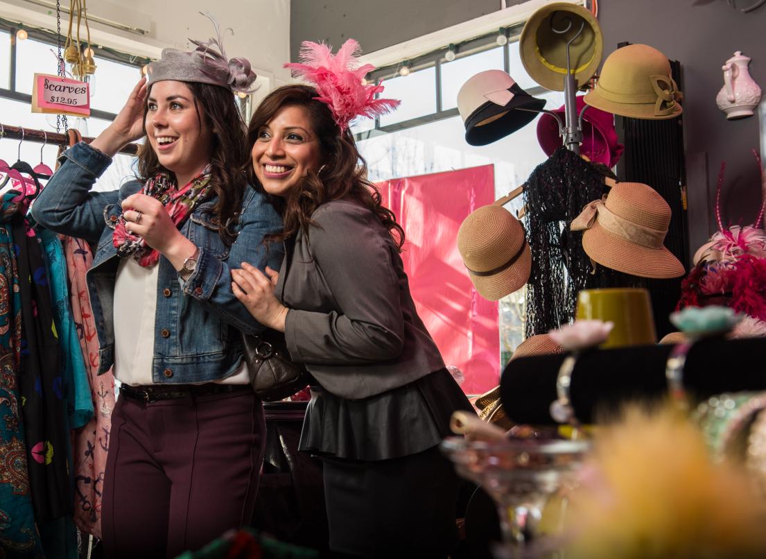 Two women smile as they try on hats at a local clothing boutique.