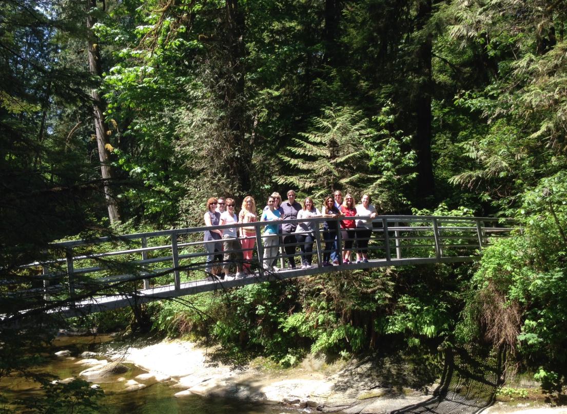 A group of people stand on a wooden bridge over a creek in the forest.