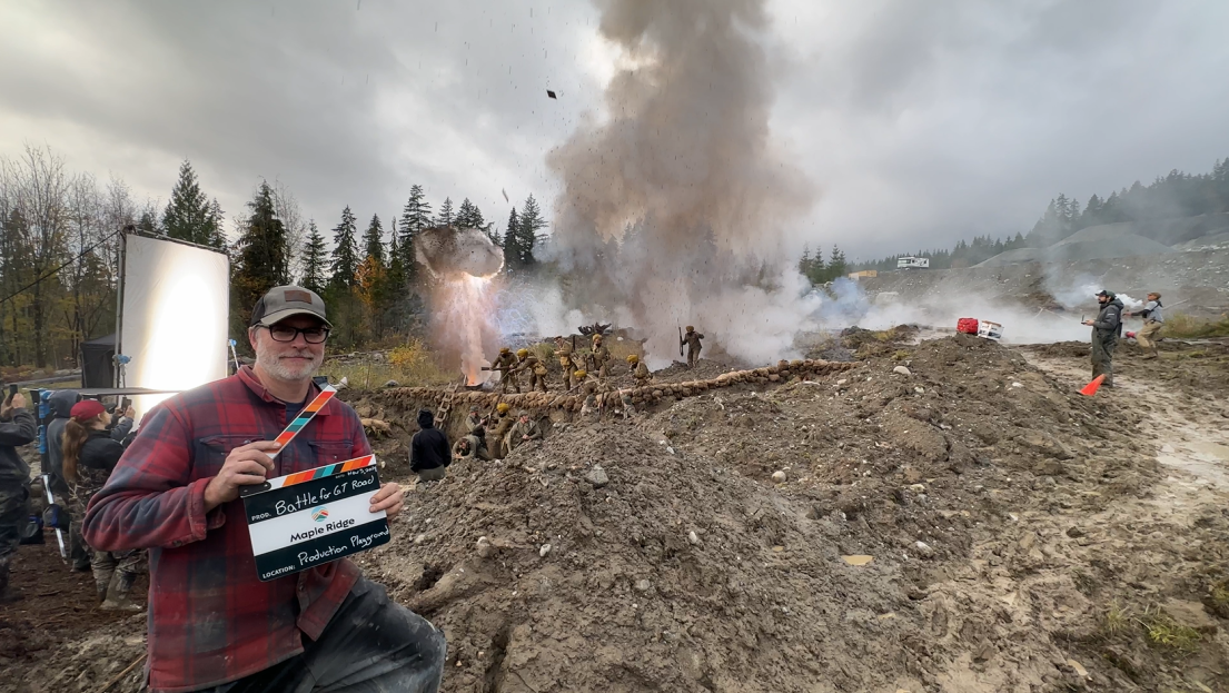 A man holding a Maple Ridge film clapper in the foreground of several explosions behind him