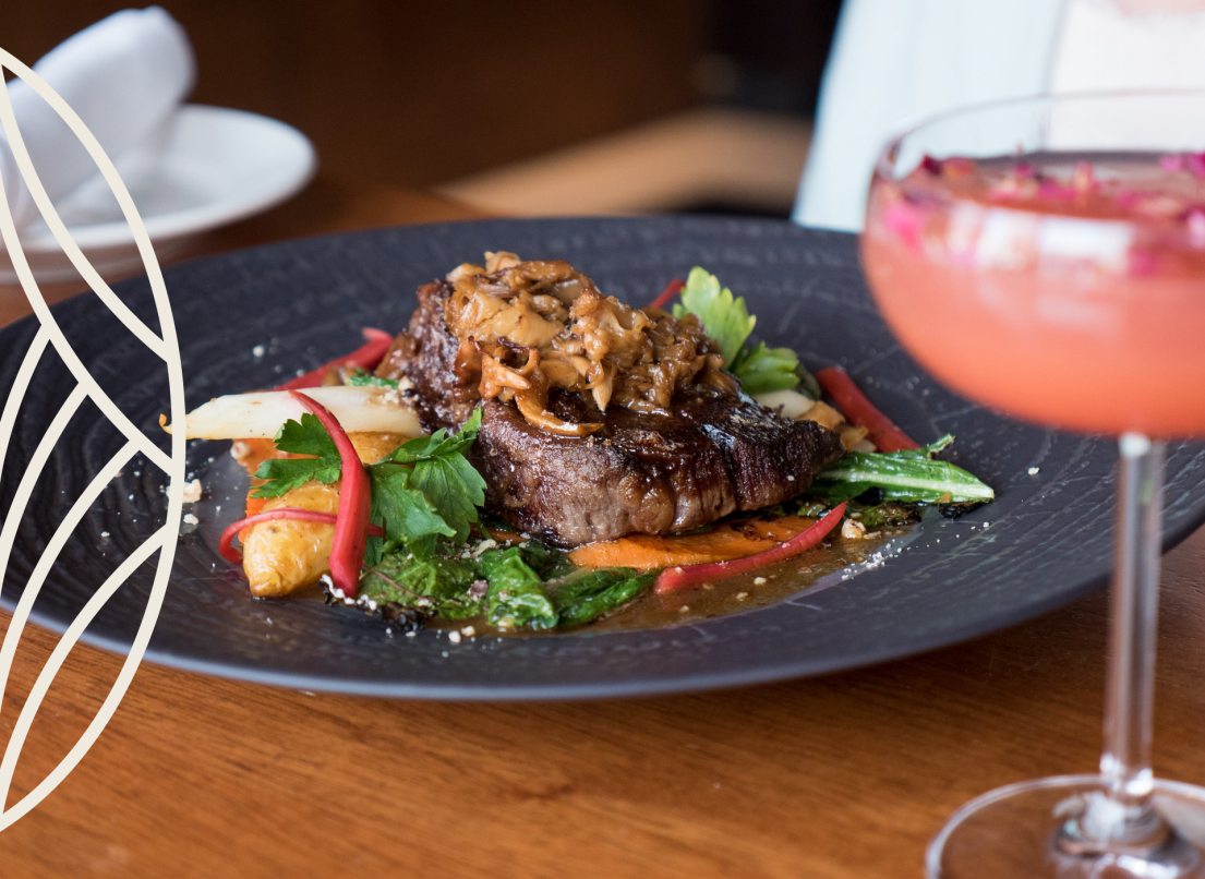 A steak rest on a bed of greens next to a champaign coupe.