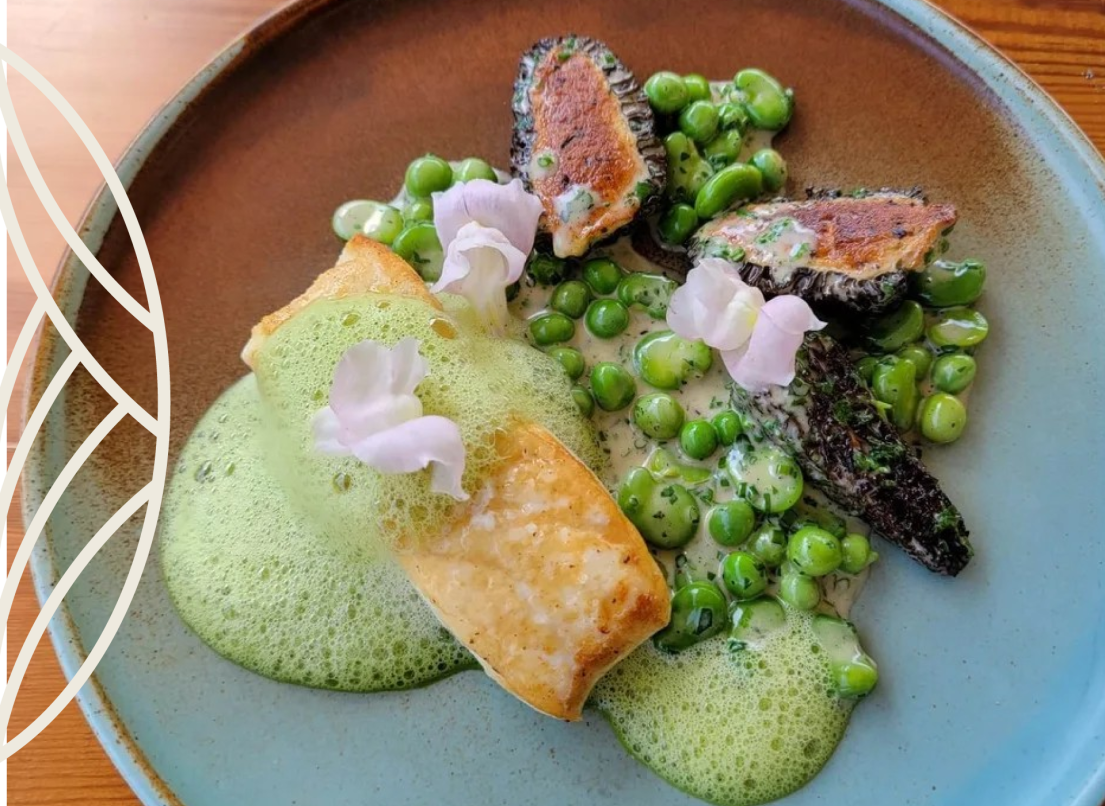 A stuffed puff pastry sits in a green sauce aside a serving of peas edible flours, and eggplant.