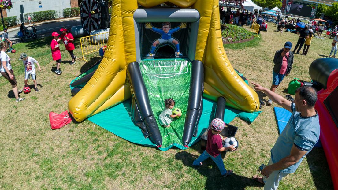 A man points two children with soccer balls towards the goal, which is up the ramp of a giant inflatable shaped like the World Cup trophy.