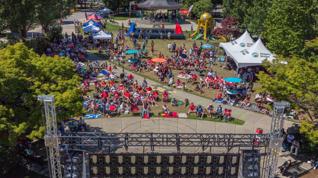 An aerial view of the crowd from behind the 20 foot tall viewing screen, showing a vibrant dotting of Canada's red-and-white colours against Memorial Peace Park's green grass.