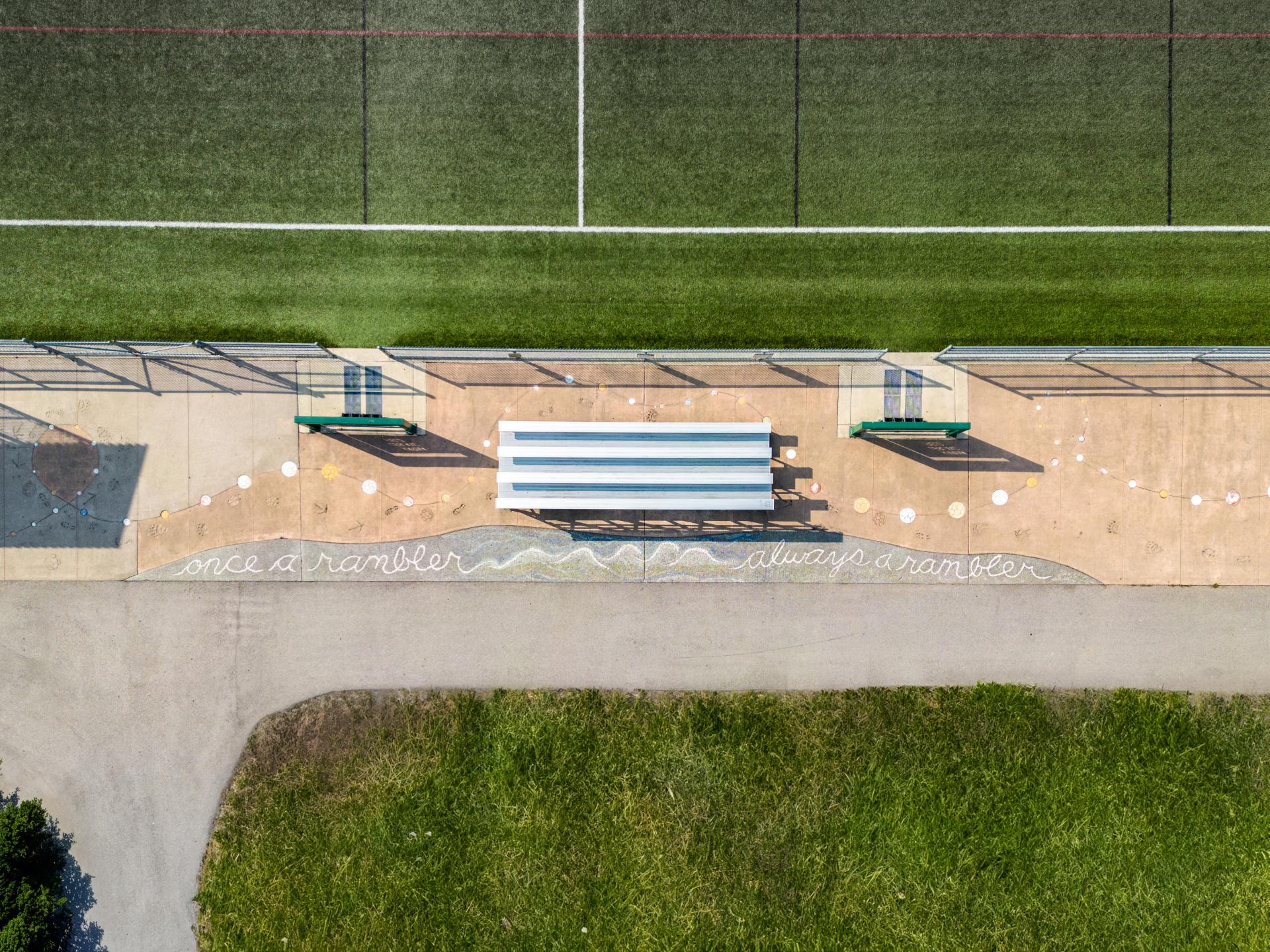 A bird's eye view of the public art installation at Karina LeBlanc Field in Merkley Park. A line of coloured soccer balls imprints are set into the concrete next to a paved pathway proclaiming "Once a rambler, always a rambler."