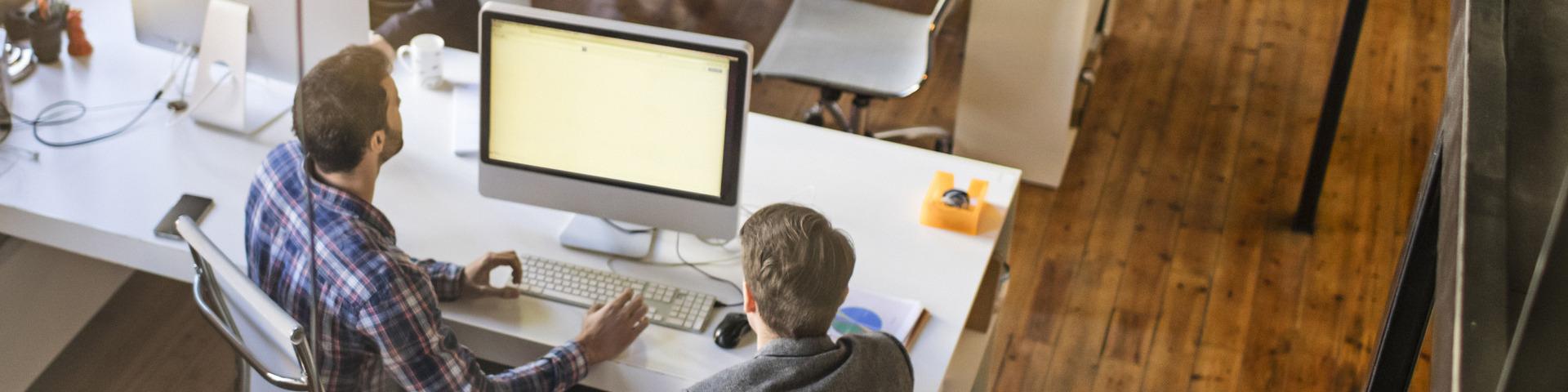 Two men review the contents of a computer screen in an office.