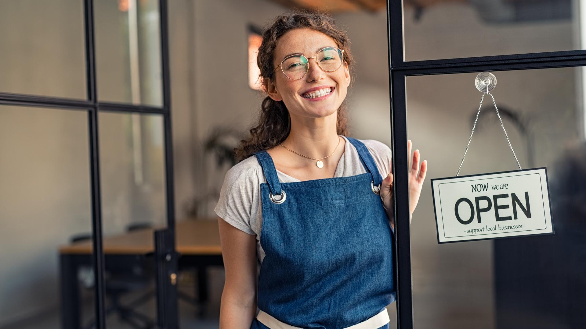 A young woman smiles as she stands in an open doorway, next to a sign welcoming customers with "Now We are Open, Support Local Businesses."