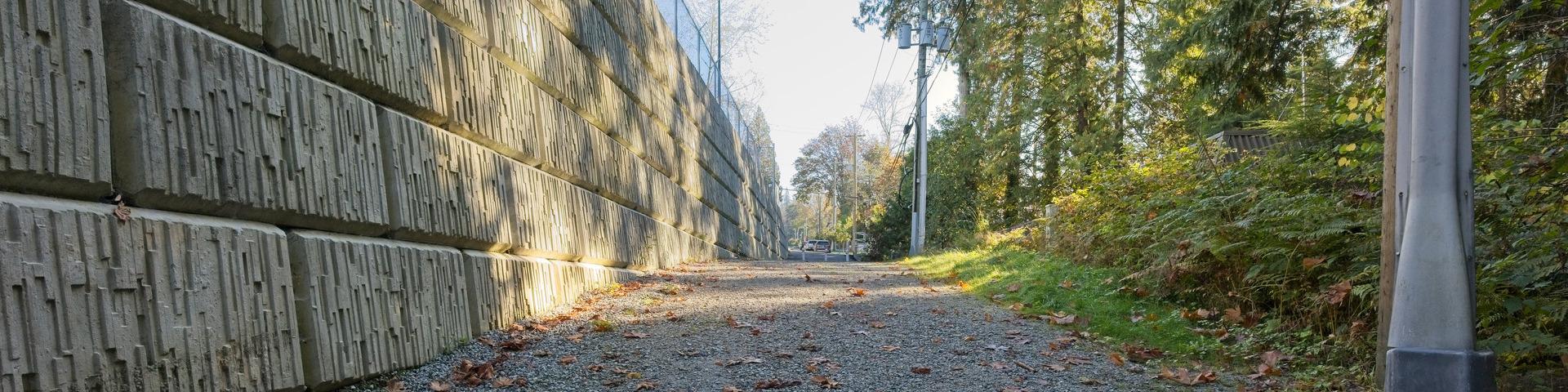 A gravel road sits between a retaining wall and a line of hydro poles.