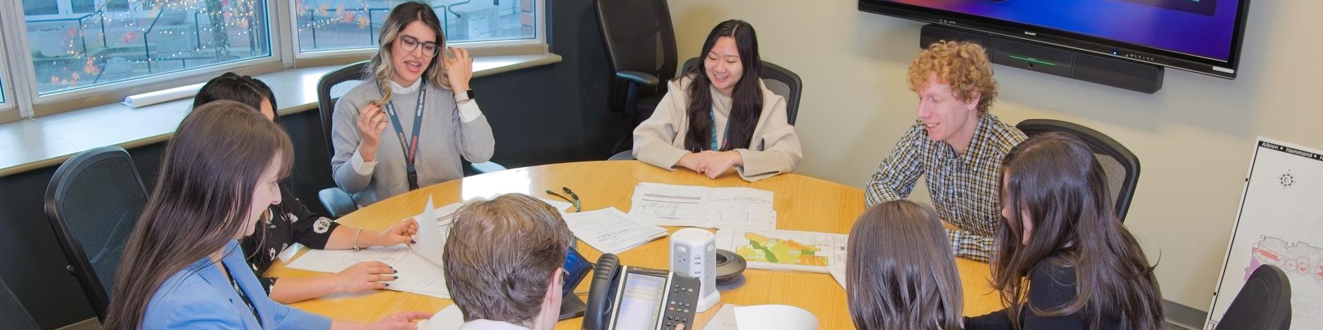 A diverse group of people sit around a table, examining multiple documents and discussing.