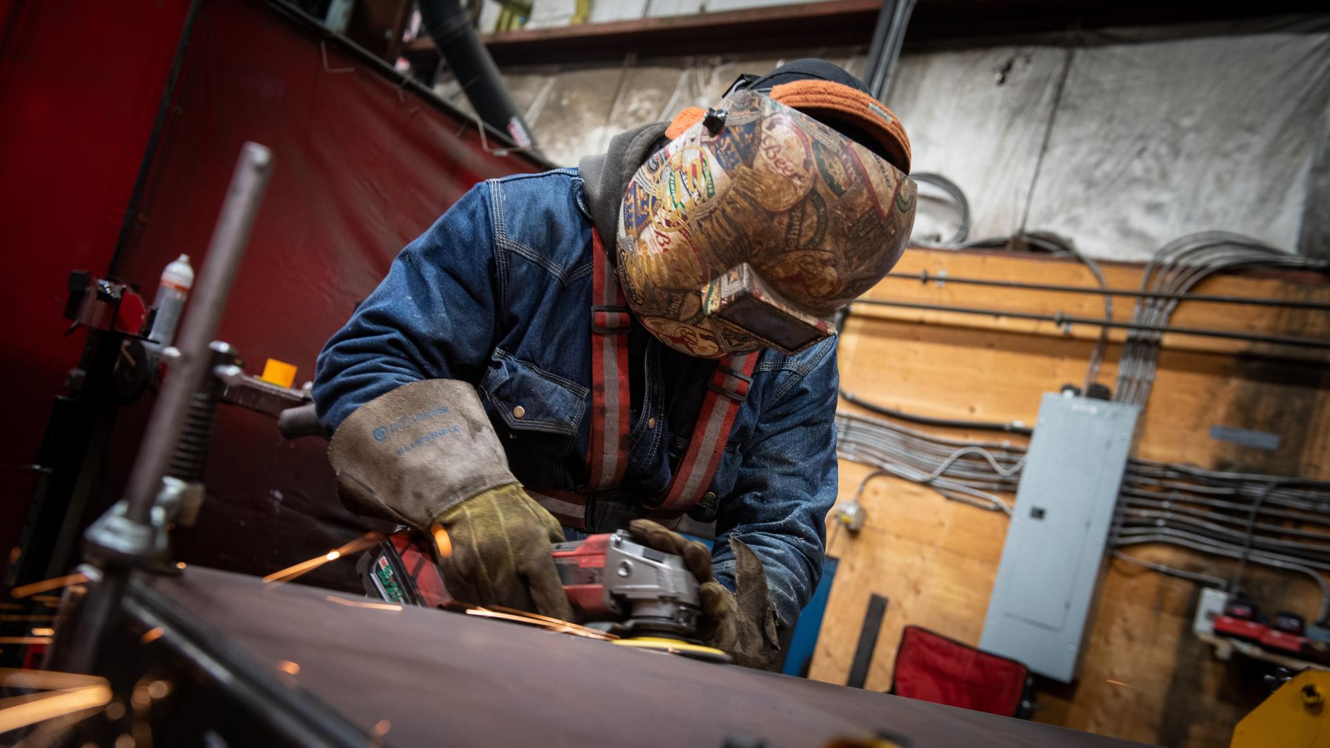 A man wearing protective equipment including a full-face helmet sands down a sheet of metal, sending sparks flying.