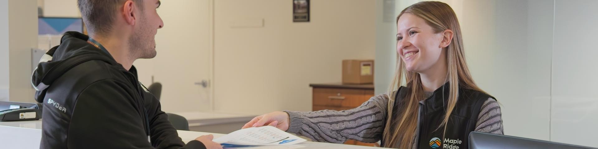 A female staff member hands a man a sheet of paper at the front desk of the Maple Ridge Leisure Centre with a wide smile on her face.