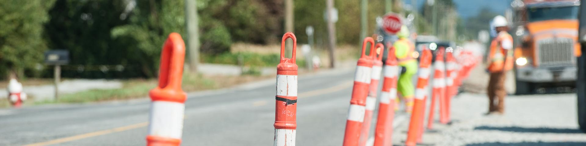 A row of orange pylons lines a roadside work area where workers in high visibility equipment are directing traffic.