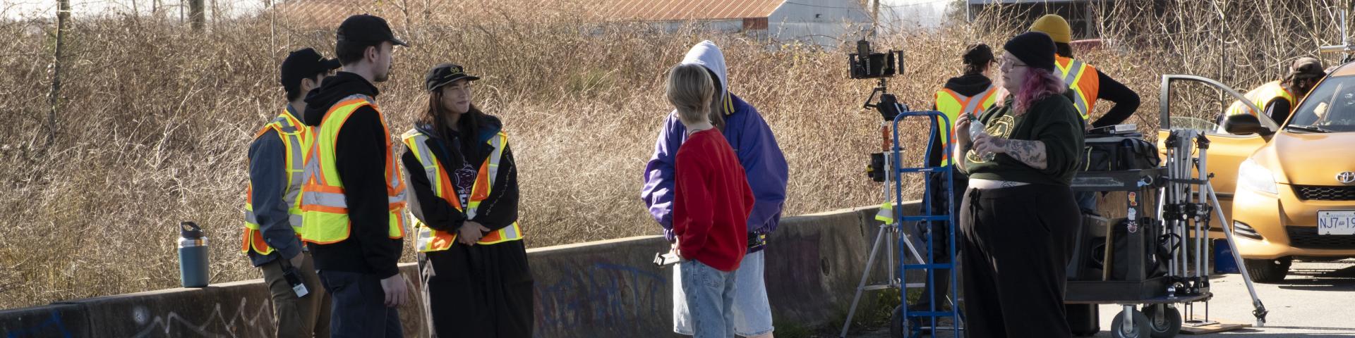 A film crew rests against a concrete barrier at the side of the road having a discussion.